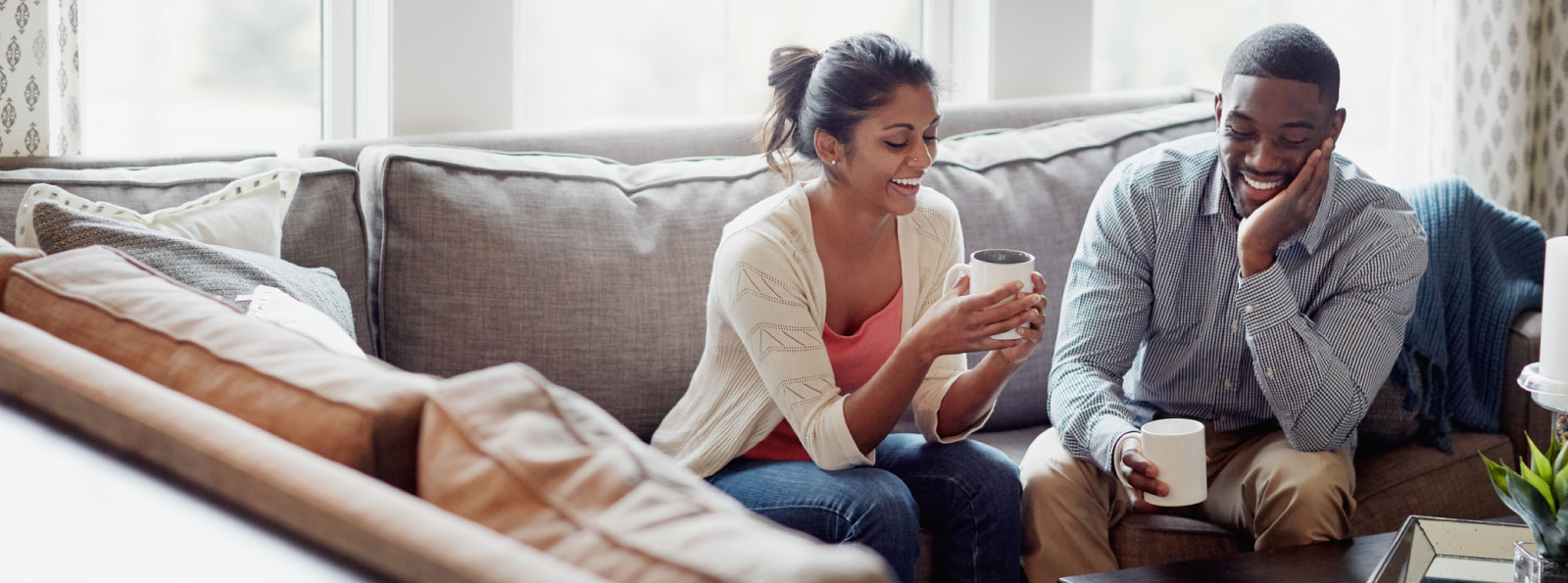 Couple sitting on couch drinking coffee and smiling at each other.